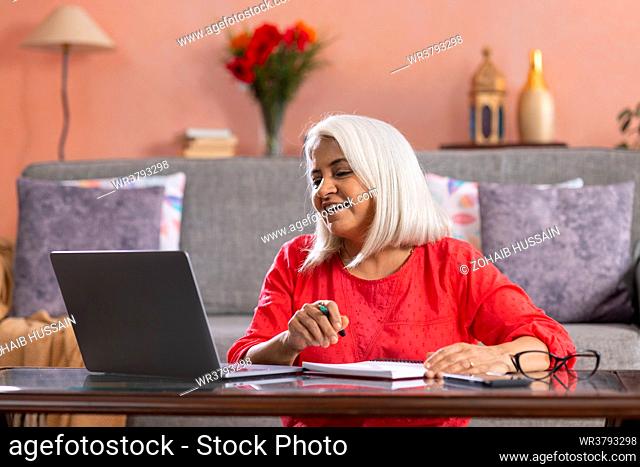 Old woman looking at laptop while writing on notepad at home