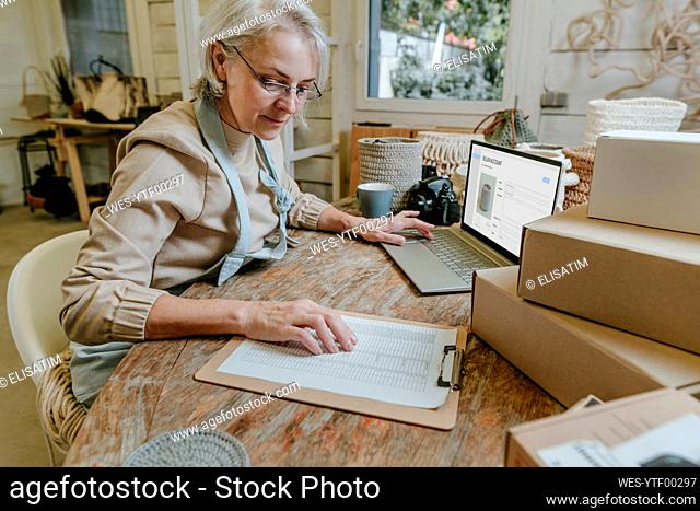 Craftswoman reading checklist sitting with laptop on workbench