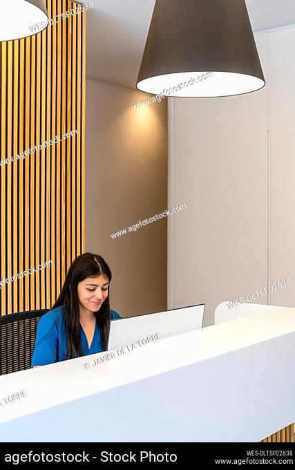 Smiling receptionist using computer at reception desk in medical clinic