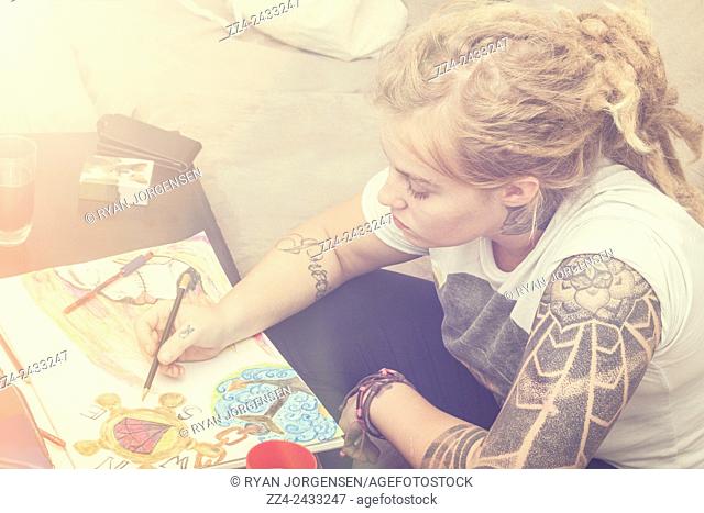 Candid photo of a beautiful blonde girl in early twenties drawing up a complex water colour sketch on padded paper. Graphic designer
