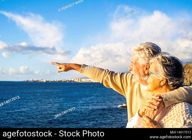 portrait of couple of mature and old people enjoying summer at the beach looking to the sea smiling and having fun together with the sunset at the background