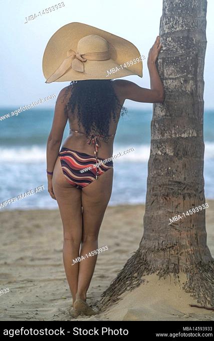 North America, Caribbean, Greater Antilles, Hispaniola Island, Dominican Republic, Puerto Plata Province, Cabarete, Attractive woman leaning against a palm tree...