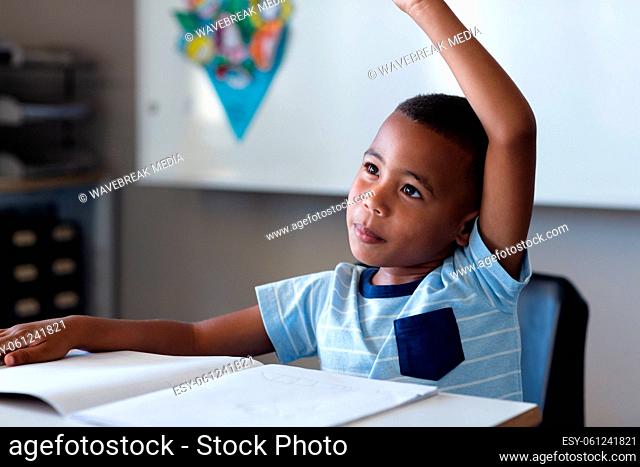 Smiling african american elementary schoolboy with hands raised sitting at desk in classroom