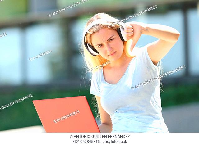Student with laptop and earbuds gesturing thumbs down outdoors in an university campus