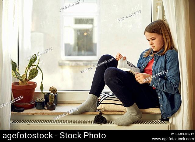 Girl sitting at the window at home reading a book