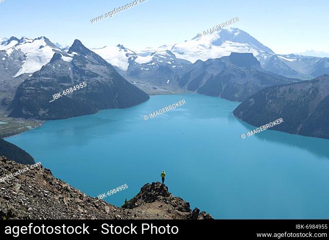 Young man standing on a rock, looking into the distance, view of mountains and glacier with turquoise blue lake Garibaldi Lake, peaks Panorama Ridge