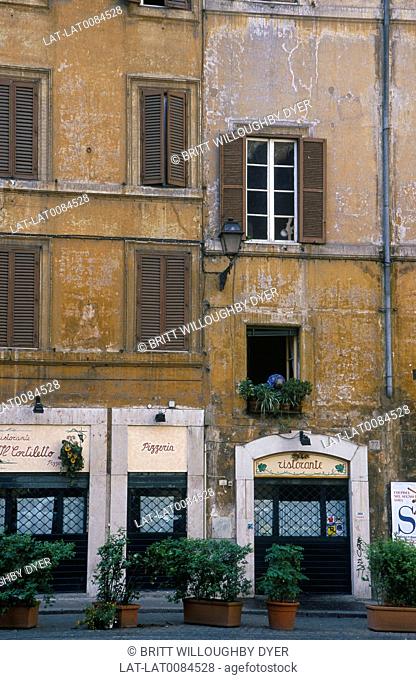 Back street. Faded brown plaster on walls. Pizzeria. Shutters closed. Person stooping to water plants