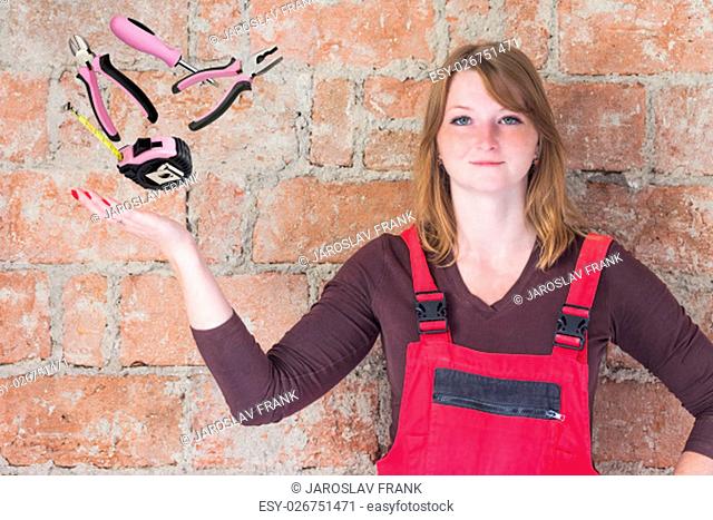 Young woman dressed in red overall is standing in front of an old brick wall. Woman is looking at the camera. Beyond her outstretched palm hand are DIY tools in...