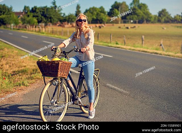 Middle-aged blond woman shopping for groceries on her bicycle stopped at the side of the road with a basketful of healthy fresh produce smiling as she faces...