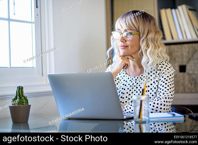 Modern web worker adult woman rest and have break relax time at the desktop in home office workstation looking outside the window and thinking