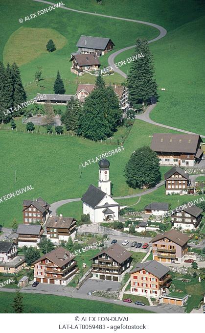 Aerial view onto alpine village in green fields. Church and tower. Chalet style houses. Road