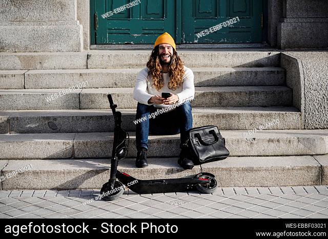 Laughing male hipster with mobile phone sitting on steps during sunny day