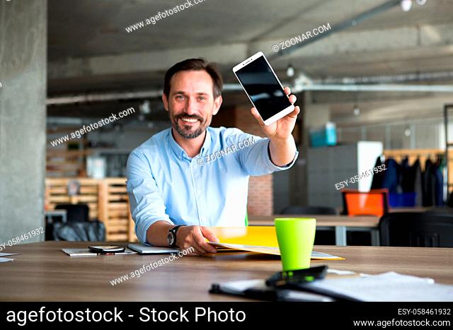 Portrait of happy smiling businessman showing mobile or smart phone with blank screen while working with documentation in office interior