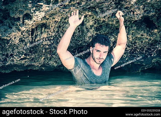 Attractive young athletic man in the sea or ocean by the rocky shore, wearing wet t-shirt, serious expression