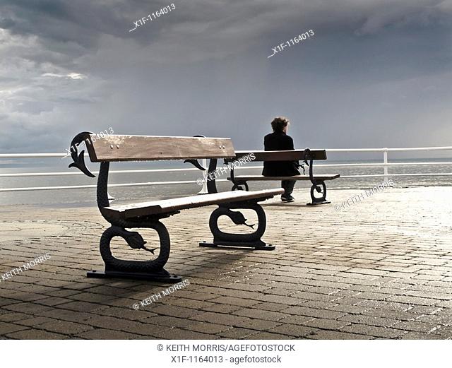 A man sitting alone on a bench on the promenade, grey overcast July summer afternoon, Aberystwyth Wales UK