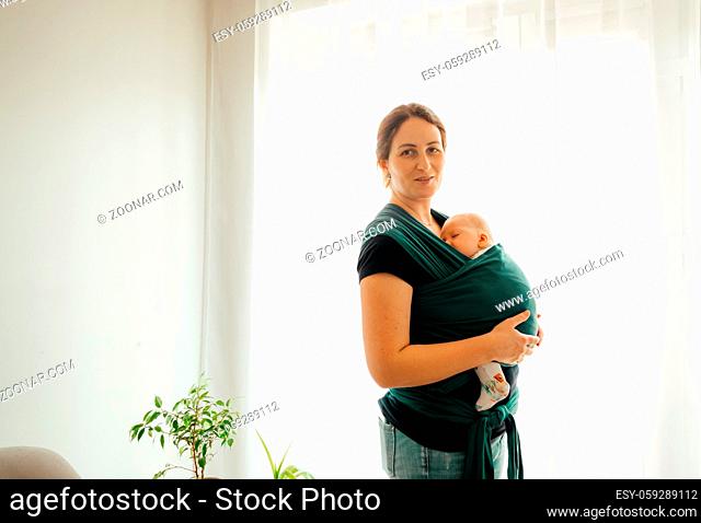 Portrait of beautiful smiling mother carrying newborn baby in dark blue baby wrap, posing next to the window at home. Cute calm baby sleeping in mother's hands