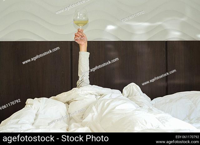 Drinking wine early tomorrow. Woman in bed, under duvet, with arm raised up holding glass of wine. Morning wine in bed