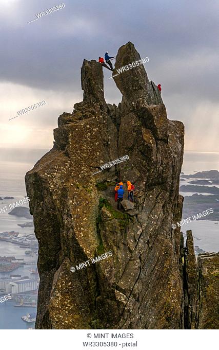 A group of people climbing on the Svolvær Goat, 150-metre (490 ft), a jagged pinnacle high above the landscape of the Lofoten