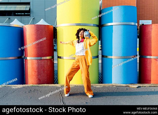 Cheerful young woman carrying boom box on shoulder dancing in front of colorful pipes