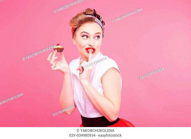 Please do not tell anyone. Portrait of beautiful young woman eating small pie secretly. She is standing and raising finger to her lips
