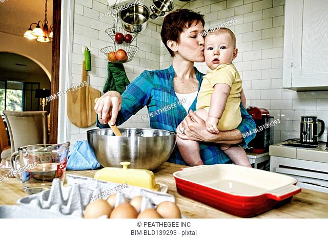 Caucasian mother kissing baby and cooking in kitchen