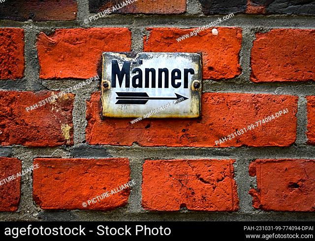 28 October 2023, Berlin: A white sign with chipped enamel and the word ""Men"" underlined by an arrow pointing to the right is affixed to a brick wall