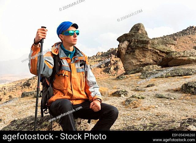 A lonely tourist with a backpack and sticks for movement in the mountains. In sunglasses and with a backpack rest sidiya on a stone
