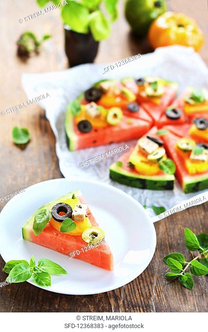 A piece of melon garnished like a pizza with tomatoes, tofu, olives and fresh herbs