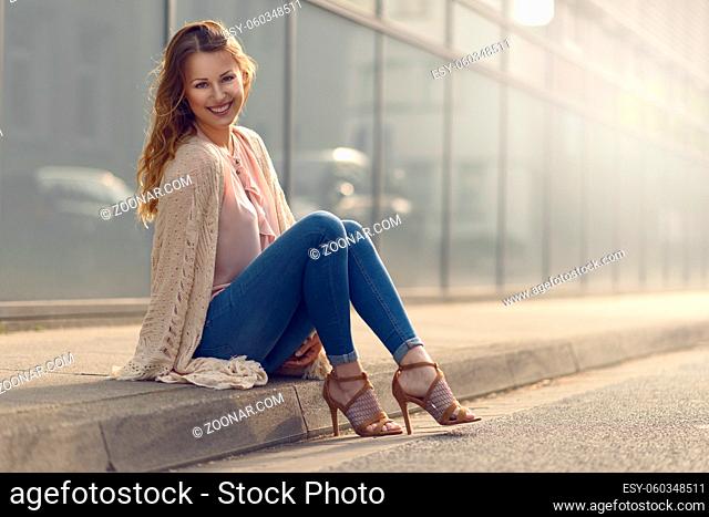 Smiling pretty trendy young woman sitting relaxing on a sidewalk on an urban street in her high heels and fashionable outfit smiling at the camera