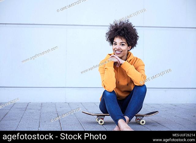 Smiling Afro woman sitting on skateboard in front of wall