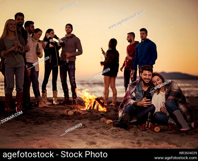Couple using cell phone during beach party with friends drinking beer and having fun colored filter