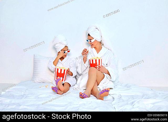 happy woman with daughter in white bathrobes and towels on heads are watching movie on tv sitting on bed