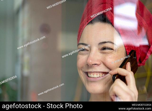 Portrait of happy woman with head wrapped in a towel talking on the phone behind windowpane
