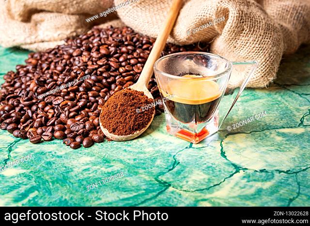 Coffee beans in coffee burlap bag on green table, wooden spoon with ground coffee on top and coffee glass cup