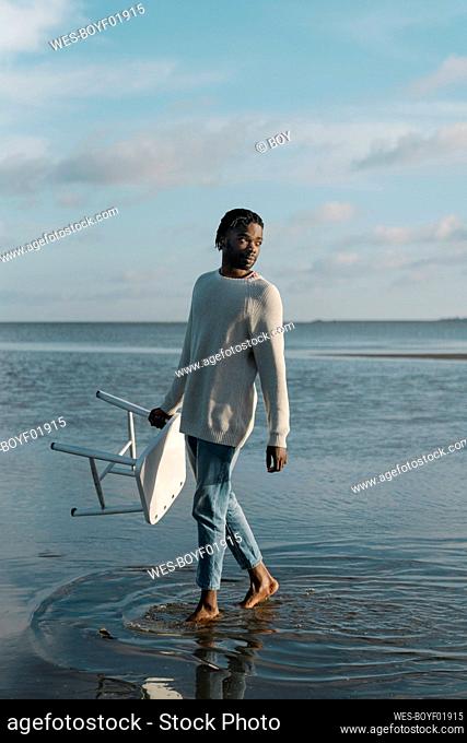 Man with white stool looking away while walking at beach against sky