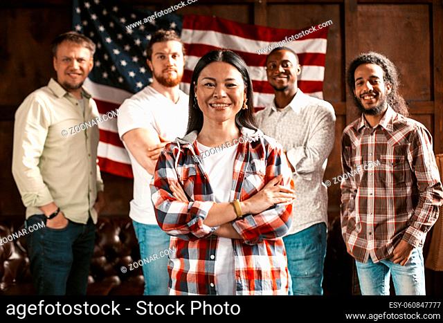 A Team Of American Patriots Against The Background Of A Wooden Wall With An American Flag. A Multi-National Group Of Young Businessmen Smiles At The Camera