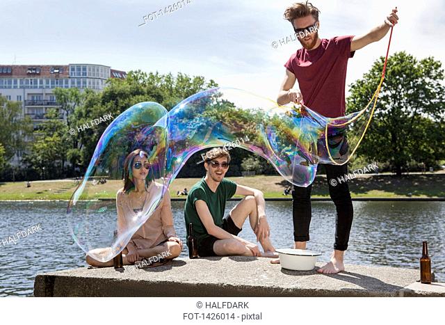 Happy friends enjoying with large bubble on retaining wall by canal at park