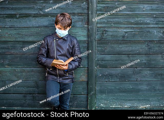 portrait of 11 year old boy with covid mask, leaning against wooden wall reading a book