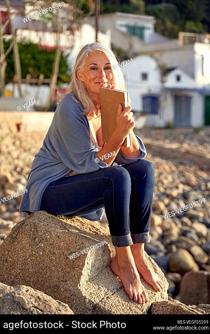 Smiling woman with book sitting on rock at beach