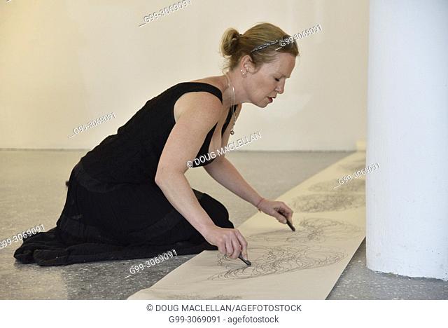 A woman artist wearing a black dress and on her knees draws with both hands as she creates a performance art work at an artist run gallery in Windsor, Canada