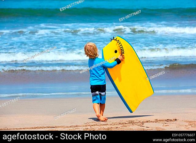 Baby boy - little surfer run with bodyboard to sea for riding on waves. Active family lifestyle, kids watersport lessons