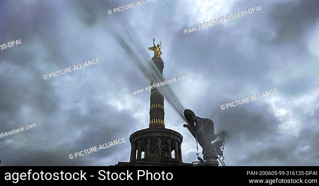 05 June 2020, Berlin: Dark clouds can be seen above the victory column, while in the foreground the water jet of a lawn sprinkler shoots into the air