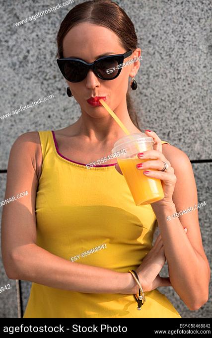 Close-up portrait of beautiful woman drinking cocktail outdoors. Girl with red lips posing in little yellow dress and sunglasses