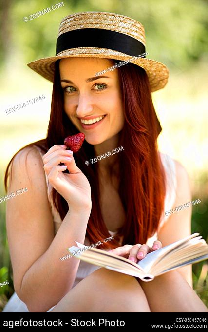 Summertime in the garden. Romantic young lady in the garden, eating strawberry and reading book