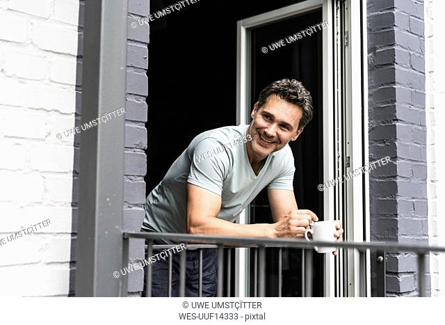 Smiling man in pyjama at home with cup of coffee looking out of balcony door
