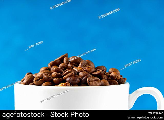 International day of coffee concept. close-up white coffee cup full of coffee beans on blue background