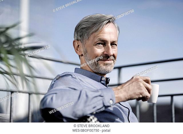 Portrait of smiling grey-haired man sitting on balcony and drinking coffee