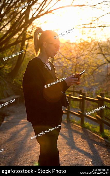 Backlit rear view of young woman talking on cell phone outdoors in park at sunset. Girl holding mobile phone, using digital device, looking at setting sun