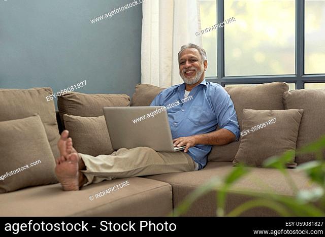 A HAPPY OLD MAN SITTING COMFORTABLY ON SOFA WITH LAPTOP