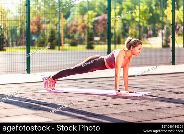 Full length, fit beautiful woman in tight pants practicing yoga on mat outdoor summer day, doing perfect plank asana, phalankasana pose and looking serious...